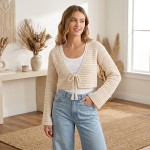 May include: A cream-colored, long-sleeved, cropped crochet cardigan featuring a front tie closure with tassels. The garment is worn over a plain white tank top and light blue denim jeans, showcasing a textured knit pattern and flared sleeves.