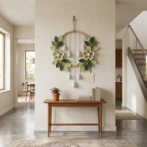 May include: A circular wall hanging featuring a wooden hoop adorned with crocheted light green flowers and dark green leaves. White yarn strands hang from the hoop, each ending in a small white teardrop-shaped ornament. A rectangular tag is attached.