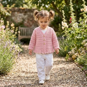 May include: A light pink crochet cardigan with wooden buttons and a scalloped hem. The cardigan is worn with white pants and shoes. The image is taken outdoors on a stone path, surrounded by flowers and greenery.