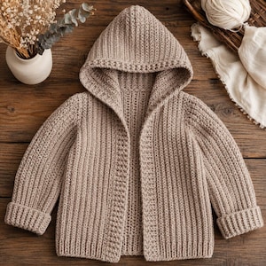 May include: A beige, hooded crochet cardigan with long sleeves. The open-front sweater features a ribbed design and folded cuffs. The hood is also ribbed. The cardigan is displayed on a wooden surface.