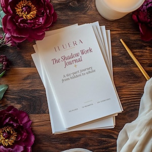 May include: A stack of white journals with the title "The Shadow Work Journal" and the text "A six-part journey from hidden to whole." The journals are on a dark wooden surface, surrounded by burgundy peonies, a lit candle, and a gold pen.