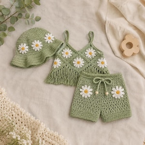May include: A sage green crochet set consisting of a bucket hat, a sleeveless crop top with fringe, and shorts. Each piece features white daisy motifs with yellow centers. The shorts include a drawstring waist with small wooden beads at the ends.