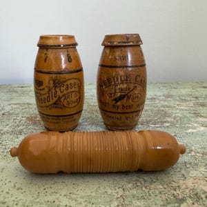 May include: Three antique wooden needle cases and a needle holder. The needle cases are barrel-shaped with printed text and numbers. The needle holder is a long, cylindrical shape with a ribbed design. The items are in shades of brown.