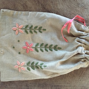 May include: A beige linen drawstring bag with pink and green embroidered flowers. The bag has a pink ribbon drawstring.