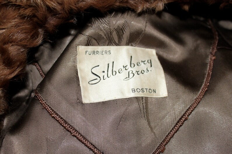 May include: A white label with black text that reads "Furriers Silberberg Bros. Boston" on a brown fur coat.