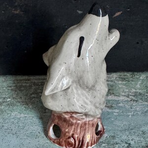 May include: A ceramic coin bank shaped like a howling wolf with a speckled light gray finish and a black nose. The wolf is perched on a brown, textured tree stump base that features two circular cutouts near the bottom and a coin slot on the neck.