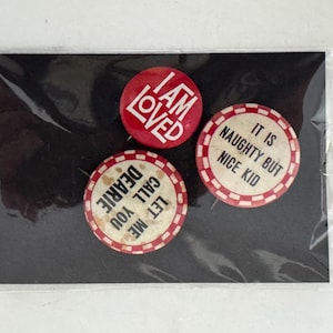 May include: Three vintage pinback buttons on a black card. One red button reads "I AM LOVED" in white. Two other buttons have a white background with red trim and text: "IT IS NAUGHTY BUT NICE KID" and "LET ME CALL YOU DEARIE".