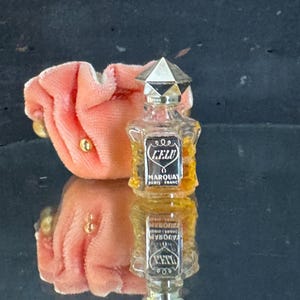 May include: A small, vintage perfume bottle with a geometric silver cap and a label that reads "LELU MARQUAY PARIS FRANCE." The bottle is next to a peach-colored velvet pouch with gold-colored accents. The bottle's reflection is visible.
