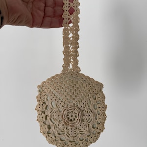 May include: A small, crocheted bag with a cream-colored lace handle. The bag is made of a delicate, open-weave pattern with a floral design in the center.