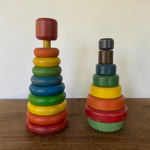 May include: Two vintage wooden stacking toys. The larger toy is a tall tower with a red top and rings in shades of red, orange, yellow, green, and blue. The smaller toy is a shorter tower with a blue top and rings in shades of blue, green, yellow, orange, and red.