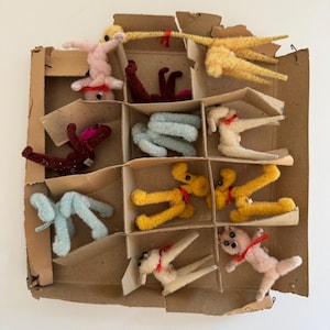 May include: A box of felt animal figurines. The box is divided into compartments and contains a variety of animals, including a pink pig, a yellow dog, a blue dog, a brown dog, and a white dog. The animals are all in different poses.