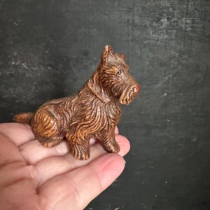 May include: A small, brown, carved dog figurine with a red nose and a collar. The dog is sitting, with its tail curled. The figurine has detailed fur texture and is likely made of ceramic or a similar material.