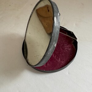 May include: A vintage, round, metal compact mirror with a hinged lid. The mirror is open and the inside is lined with a dark red fabric. The mirror has a silver finish and shows signs of wear.