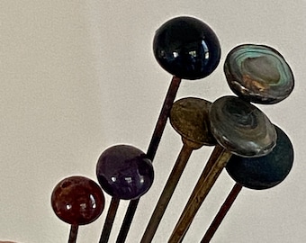 May include: A set of seven vintage glass-headed hat pins. The pins are made of metal and have different colored glass heads, including black, purple, brown, green, and gray.