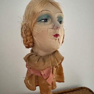 May include: A vintage doll head with blonde hair, blue eyeshadow, red lips, and a brown fabric collar. The doll's face is cracked and shows signs of age.