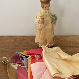 May include: A small porcelain figurine of a child wearing a gold crown and a white robe, holding a blue and gold orb. The figurine is surrounded by a stack of colorful fabric scraps.
