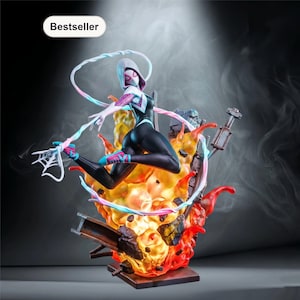 May include: A dynamic figurine of a superheroine in a black and white suit, leaping from a fiery base. The figure has a white hood and mask, with a web-slinging pose. The base features flames and industrial elements, with a "Bestseller" label.