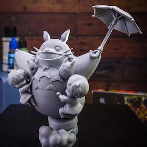 May include: A gray figurine of Totoro holding an umbrella, with three small figures clinging to its body. The figurine is on a cloud-shaped base. The background is a wooden surface.