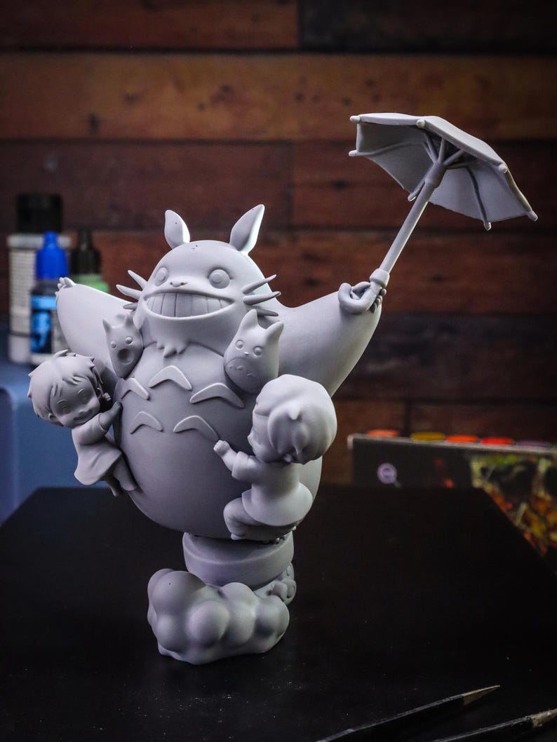 May include: A gray figurine of Totoro holding an umbrella, with three small figures clinging to its body. The figurine is on a cloud-shaped base. The background is a wooden surface.
