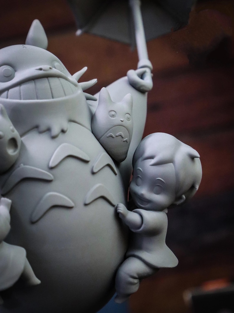 May include: A gray figurine depicting characters from a popular animated film. The central figure is a large, smiling creature with a round body, holding an umbrella. A smaller character clings to its side, and other figures are also present.