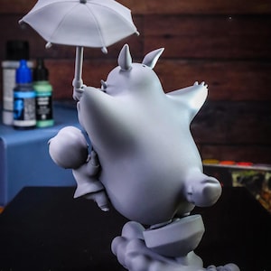 May include: A gray figurine of a large, round creature holding an umbrella, with a smaller figure beside it. The creature has pointed ears and is standing on a cloud-like base. The umbrella is open, and the overall design is whimsical.