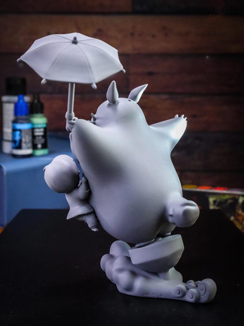 May include: A gray figurine of a large, round creature holding an umbrella, with a smaller figure beside it. The creature has pointed ears and is standing on a cloud-like base. The umbrella is open, and the overall design is whimsical.