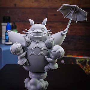 May include: A gray figurine of Totoro holding an umbrella, with two children and two smaller Totoros. The figurine is on a cloud-shaped base. The background is a wooden wall.
