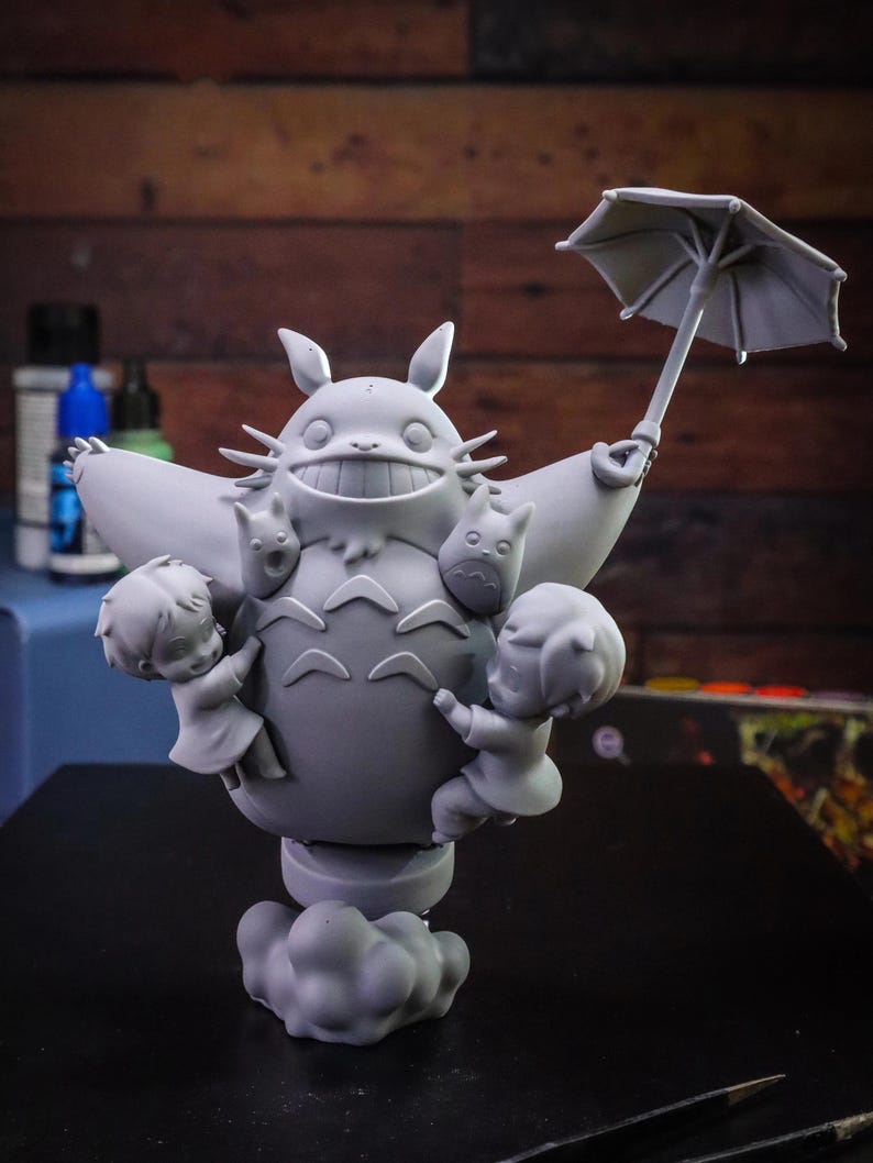May include: A gray figurine of Totoro holding an umbrella, with two children and two smaller Totoros. The figurine is on a cloud-shaped base. The background is a wooden wall.