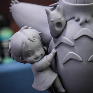 May include: A gray figurine depicting a scene from an animated film. A smiling child figure is clinging to a large, round, furry creature. A smaller, cat-like figure is attached to the larger creature.