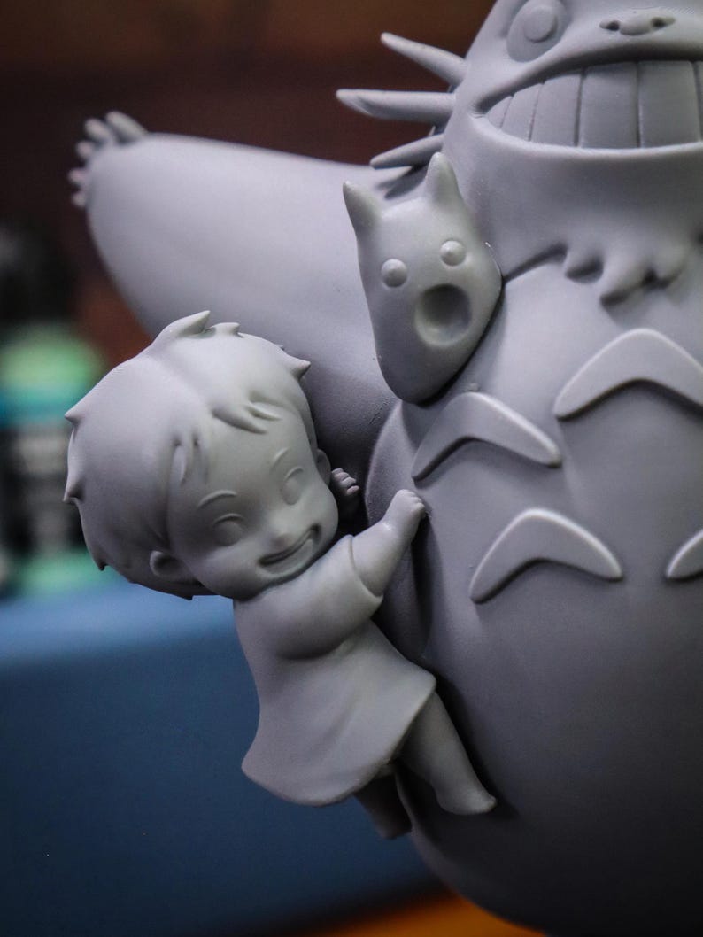 May include: A gray figurine depicting a scene from an animated film. A smiling child figure is clinging to a large, round, furry creature. A smaller, cat-like figure is attached to the larger creature.