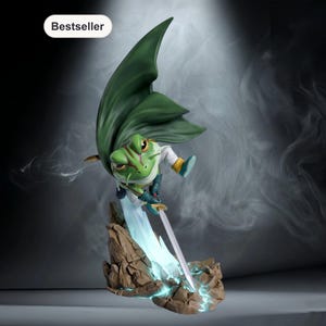 May include: A green frog figurine with a cape and sword, standing on a rocky base. The frog is wearing a white shirt and blue boots. The word "Bestseller" is in a white oval in the upper left corner.