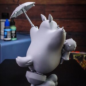 May include: A gray figurine of a cartoon character holding a small umbrella. The character has large ears and is standing on a cloud-like base. A smaller figure is held in its arms. The figurine is set against a blurred background.
