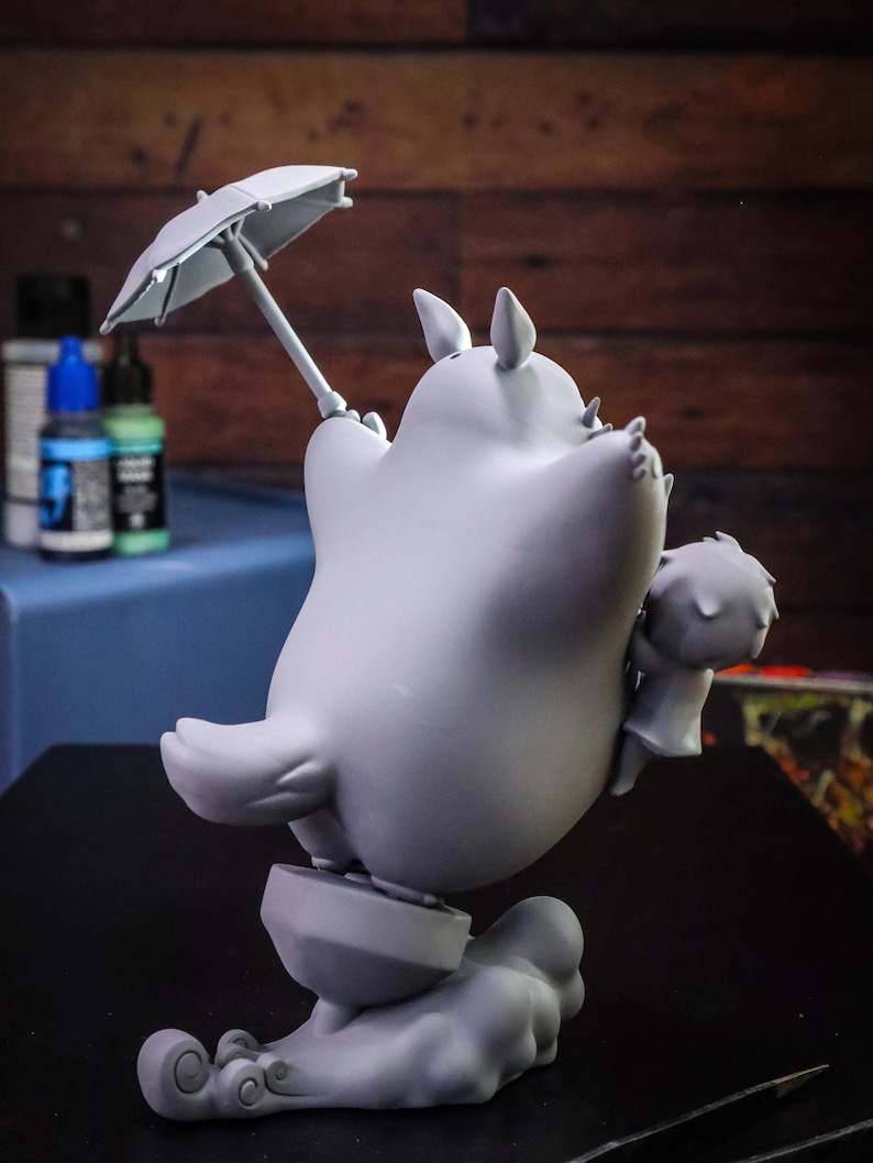 May include: A gray figurine of a cartoon character holding a small umbrella. The character has large ears and is standing on a cloud-like base. A smaller figure is held in its arms. The figurine is set against a blurred background.