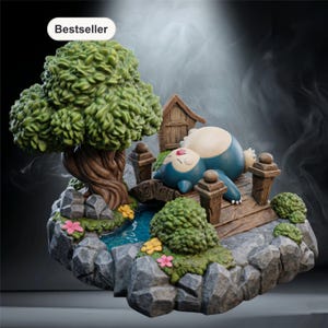 Op de afbeelding: Gedetailleerd beeldje van een slapend blauw en crèmekleurig wezen op een houten brug, met een klein huisje en een grote groene boom. De scène omvat een kleine vijver, rotsen en bloemen, waardoor een miniatuurlandschap ontstaat. Het woord "Bestseller" staat linksboven.