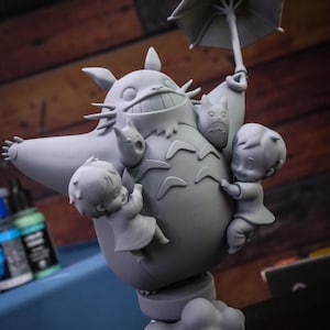 May include: A gray figurine of Totoro holding an umbrella, with two children and two smaller Totoros clinging to it. The figurine is on a base of clouds.