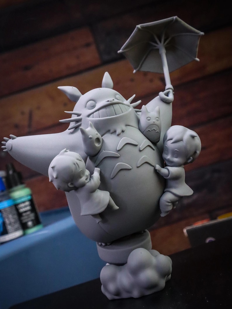 May include: A gray figurine of Totoro holding an umbrella, with two children and two smaller Totoros clinging to it. The figurine is on a base of clouds.