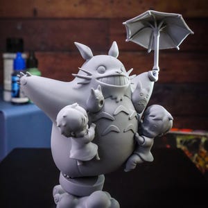 May include: A gray figurine of Totoro, a large forest spirit, holding an umbrella and surrounded by three small figures. The figurine is on a cloud-like base, with details like whiskers and a wide grin.