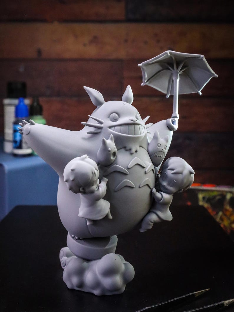 May include: A gray figurine of Totoro, a large forest spirit, holding an umbrella and surrounded by three small figures. The figurine is on a cloud-like base, with details like whiskers and a wide grin.