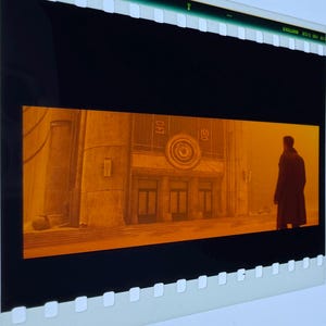 以下が含まれることがあります： モノクロームのオレンジ色で表現された映画のワンシーンが映る35mmフィルムストリップのフレーム。長いダークコートを着た人物が、円形のエンブレムと3つの出入り口がある大きな建物の前に立っている。フィルムストリップには黒い縁とパーフォレーションがある。
