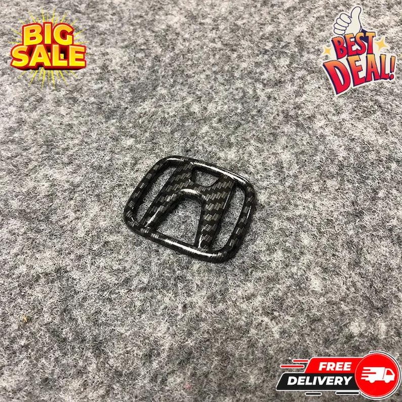 May include: A carbon fiber-style Honda emblem, featuring a black and gray weave pattern. The emblem is rectangular with rounded corners and the Honda "H" logo in the center. The background is a gray, textured surface.