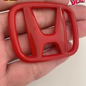 May include: A glossy red Honda emblem. The emblem is in the shape of a square with rounded corners and features the Honda logo in the center. The image also includes text that says "BIG SALE", "BEST DEAL!", and "FREE DELIVERY".
