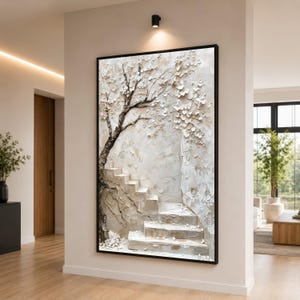 以下が含まれることがあります： 白い花と階段のある木を描いた、額入りのテクスチャアート。木の幹は濃い茶色で、階段は薄いベージュ色です。アートワークは、明るい廊下の白い壁に展示されています。