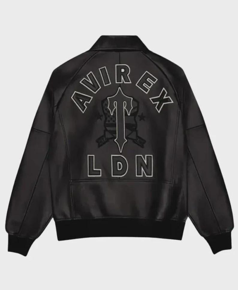 Pode incluir: Casaco de couro preto com gola e punhos canelados. As costas apresentam um design bordado branco com o texto "AVIREX" acima de um emblema estilizado e "LDN" abaixo.