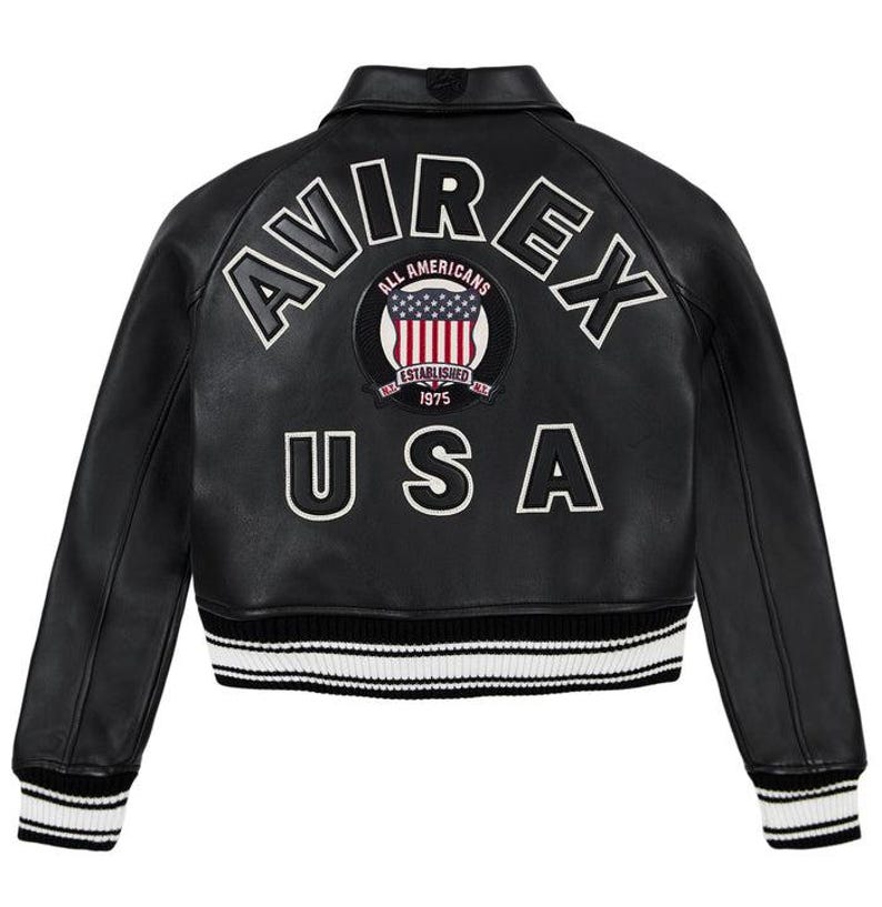 May include: Black leather jacket with white embroidered lettering spelling "AVIREX" and "USA". A circular patch with the American flag and the words "ALL AMERICANS ESTABLISHED 1975" is centered on the back. The jacket has a ribbed collar and cuffs.