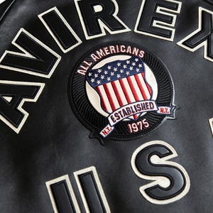 May include: Close-up of a black leather jacket with white lettering spelling "AVIREX" and "US". A circular patch features a shield with the American flag and the text "ALL AMERICANS ESTABLISHED 1975 N.Y."