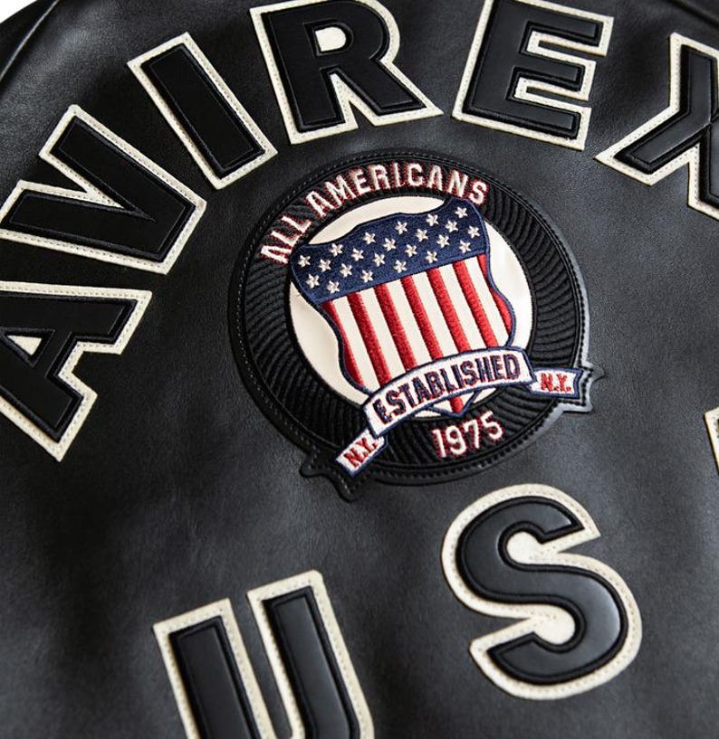May include: Close-up of a black leather jacket with white lettering spelling "AVIREX" and "US". A circular patch features a shield with the American flag and the text "ALL AMERICANS ESTABLISHED 1975 N.Y."