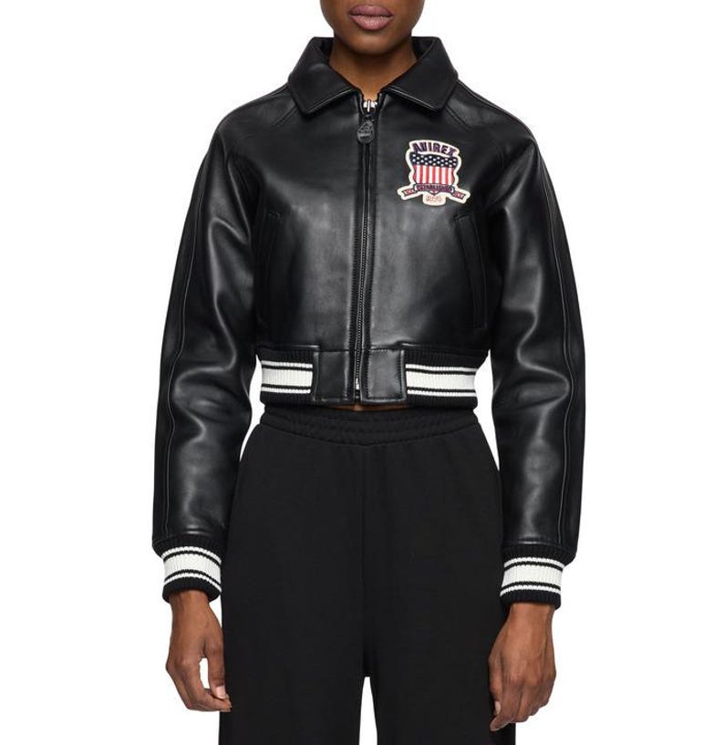 May include: Black leather bomber jacket with a cropped fit. The jacket features a white and black striped hem and cuffs. A patch with the text "AVIREX" and a patriotic design is on the chest.