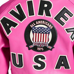 K&ouml;nnte beinhalten: Eine leuchtend pinkfarbene Jacke mit schwarzen und wei&szlig;en Buchstaben, die "AVIRE USA" buchstabieren. Ein kreisf&ouml;rmiger Aufn&auml;her zeigt ein Schild mit der amerikanischen Flagge und den Worten "ALL AMERICANS ESTABLISHED 1975".