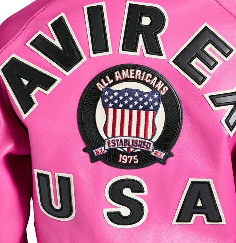 K&ouml;nnte beinhalten: Eine leuchtend pinkfarbene Jacke mit schwarzen und wei&szlig;en Buchstaben, die "AVIRE USA" buchstabieren. Ein kreisf&ouml;rmiger Aufn&auml;her zeigt ein Schild mit der amerikanischen Flagge und den Worten "ALL AMERICANS ESTABLISHED 1975".