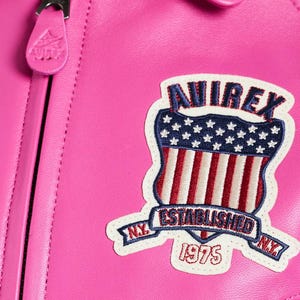K&ouml;nnte beinhalten: Nahaufnahme einer leuchtend rosa Lederjacke. Die Jacke hat einen Aufn&auml;her mit dem Text "AVIREX" &uuml;ber einem Schilddesign mit der amerikanischen Flagge und den Worten "ESTABLISHED 1975 N.Y."